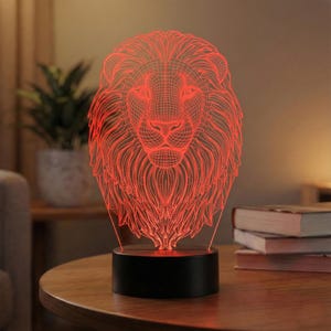May include: A red, 3D-effect lion head lamp. The lion's mane and facial features are outlined with red light, and the lamp sits on a black base. The lamp is on a wooden table.