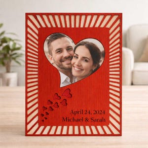 Puede incluir: Un marco de fotos de madera roja con un recorte en forma de corazón que contiene una foto de una pareja sonriente. El marco tiene un borde decorativo y el texto "April 24, 2024 Michael & Sarah".