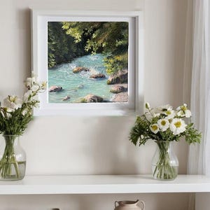 May include: A framed painting depicting a turquoise river in a green forest. Two vases of white flowers are on a white shelf beneath the artwork. The scene is lit by natural light, creating a serene atmosphere.