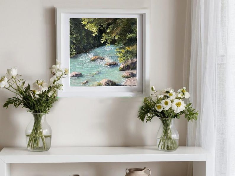 May include: A framed painting depicting a turquoise river in a green forest. Two vases of white flowers are on a white shelf beneath the artwork. The scene is lit by natural light, creating a serene atmosphere.