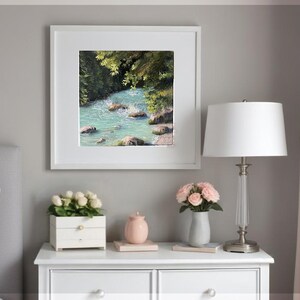 May include: A framed painting depicting a turquoise river winding through a green forest. The artwork is presented in a white frame, showcasing the natural beauty of the landscape. The painting is displayed on a white bedside table.
