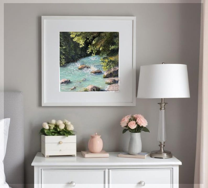 May include: A framed painting depicting a turquoise river winding through a green forest. The artwork is presented in a white frame, showcasing the natural beauty of the landscape. The painting is displayed on a white bedside table.