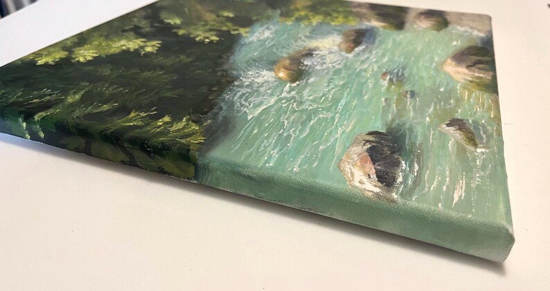 May include: A canvas painting of a turquoise river scene with rocks and green foliage. The artwork is viewed from a side angle, revealing the textured brushstrokes and the canvas edge. The painting uses metric measurements.
