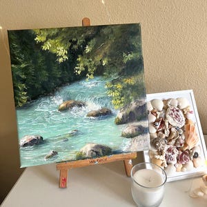 May include: An oil painting on a small wooden easel shows a turquoise river flowing over rocks, framed by green trees. A display of seashells in a white frame and a lit candle in a glass holder are also visible.