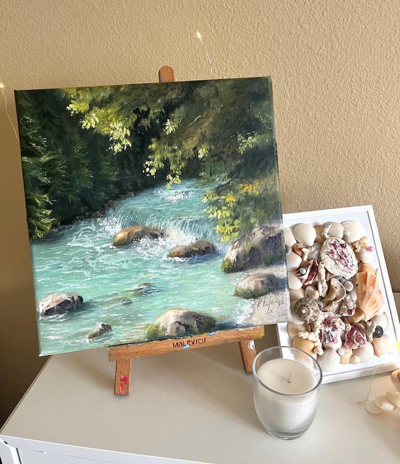 May include: An oil painting on a small wooden easel shows a turquoise river flowing over rocks, framed by green trees. A display of seashells in a white frame and a lit candle in a glass holder are also visible.