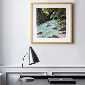 May include: A framed landscape painting shows a turquoise river in a forest setting. The artwork, in a gold frame, features rocks in the water and green foliage. A silver desk lamp and a laptop are in the foreground.