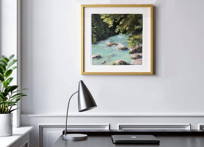 May include: A framed landscape painting shows a turquoise river in a forest setting. The artwork, in a gold frame, features rocks in the water and green foliage. A silver desk lamp and a laptop are in the foreground.