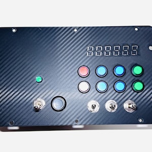 May include: A black control panel with a carbon fiber texture. It features a digital display, multiple colored buttons, toggle switches, and a green indicator light. The panel has mounting holes.