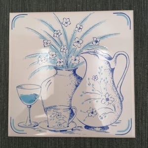 Op de afbeelding: Een vierkante keramische tegel met een blauw-witte illustratie van een kan, een theekopje op een schoteltje en een glas op voet. Een boeket bloemen staat in een vaas achter het kopje, alles in blauwe lijntekeningen op een witte ondergrond.