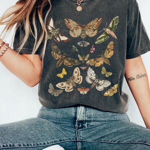 May include: A dark gray t-shirt with a vintage-style print of various butterflies and moths in shades of brown, yellow, white, and green. The shirt has a relaxed fit and short sleeves.