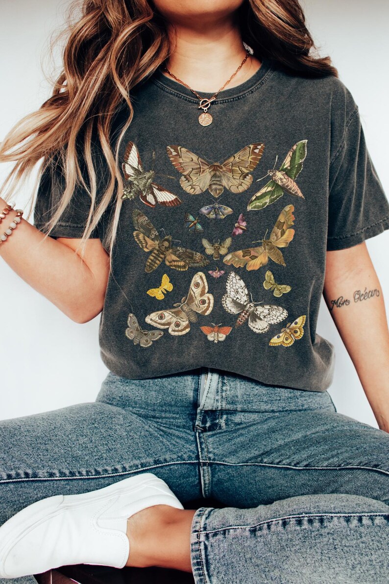 May include: A dark gray t-shirt with a vintage-style print of various butterflies and moths in shades of brown, yellow, white, and green. The shirt has a relaxed fit and short sleeves.