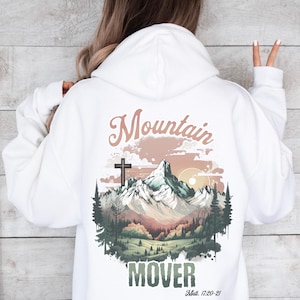 May include: White hoodie with a graphic of a mountain range with a cross at the top, a sunset, and trees. The text "Mountain Mover" is below the graphic, and the text "Matt. 17:20-21" is below that.