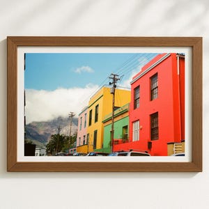 Peut inclure: Une impression encadrée représentant une scène de rue avec des bâtiments colorés. Les bâtiments sont peints dans des teintes vives de rouge, jaune, vert et rose. Une chaîne de montagnes est visible en arrière-plan sous un ciel bleu nuageux. Le cadre est de couleur marron chaud.