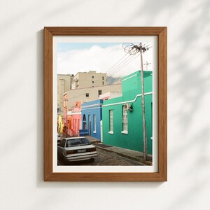 Può includere: Una stampa incorniciata con una scena di strada colorata. Gli edifici sono dipinti in vivaci tonalità di blu, verde, rosa e giallo. Un'auto bianca è parcheggiata sulla strada acciottolata. Il cielo è nuvoloso.