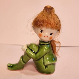 May include: A ceramic figurine of a seated elf in a green outfit and with red hair. The elf has a smiling face with rosy cheeks and is posed with its knees bent. The figurine is a decorative item.