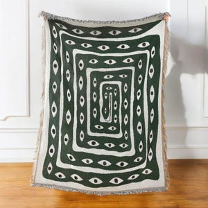 May include: A green and white woven throw blanket with a repeating pattern of eyes. The eyes are green with black pupils and are arranged in a concentric square pattern.