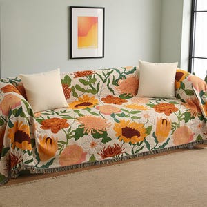 Puede incluir: Una funda de sofá con estampado floral en tonos naranja, amarillo y rosa sobre un fondo crema. Dos cojines cuadrados color crema descansan sobre el sofá. La funda tiene un borde con flecos.