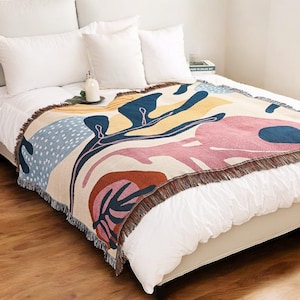 May include: A white bed with a white and cream throw blanket with a colorful abstract pattern. The blanket has a fringe border and features shades of blue, pink, yellow, orange, and brown.