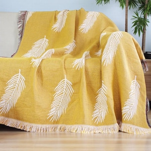 Könnte beinhalten: Senfgelbe Decke mit weißem Federmuster. Die Decke hat einen Fransenrand und ist über ein Sofa drapiert. Der Überwurf ist ein dekoratives Wohnaccessoire.