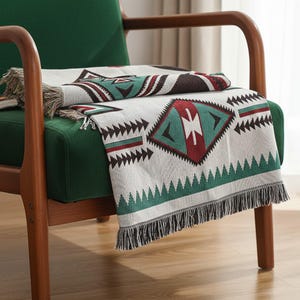 Puede incluir: Una manta tejida con un patrón geométrico en blanco, verde, rojo y marrón descansa sobre una silla verde. La manta presenta un diseño de diamantes y triángulos con flecos en los bordes. La silla tiene un marco de madera.