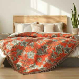 Può includere: Una coperta arancione con un motivo floreale blu e marrone copre un letto. La coperta ha un bordo con frange ed è drappeggiata su lenzuola bianche. Una testiera in legno e cuscini sono dietro la coperta.