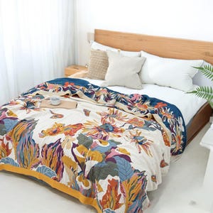 Op de afbeelding: Een bloemmotief quilt op een bed. De quilt heeft grote, kleurrijke bloemen in tinten oranje, geel, blauw en paars tegen een crèmekleurige achtergrond. De rand is donkerblauw met een gele rand. Het bed heeft witte kussens en een houten hoofdbord.