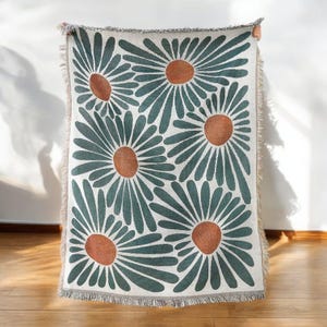 May include: A green and white throw blanket with a repeating pattern of stylised orange flowers. The blanket has a fringed edge.
