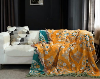 May include: A yellow and teal throw blanket with a floral pattern and fringe. The blanket is draped over a white sofa with a geometric patterned cushion.