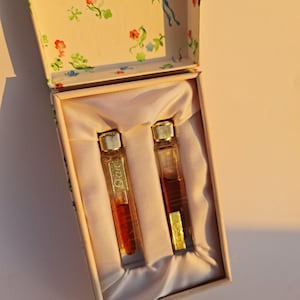 Könnte beinhalten: Zwei kleine Glasflaschen mit goldenen Kappen und brauner Flüssigkeit darin, die in einer weißen Schachtel mit einem Blumenmuster auf dem Deckel liegen.