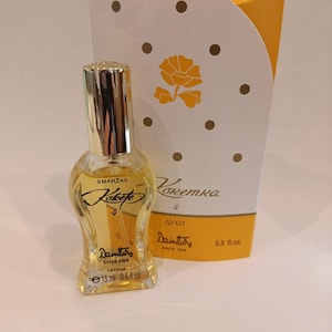 Puede incluir: Un pequeño frasco de vidrio de perfume llamado "Koketka" de Daintary, desde 1849. El frasco es amarillo y tiene una tapa dorada. La etiqueta dice "Smarzas" y "Koketka" en una fuente cursiva. El frasco está sentado en una caja blanca y amarilla con un diseño de flores.