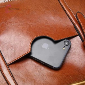 Może przedstawiać: Zbliżenie brązowej sk&oacute;rzanej torebki z kieszenią na czarny smartfon. Torba ma teksturowaną powierzchnię i widoczne szwy. Logo TotyBags znajduje się w lewym g&oacute;rnym rogu.