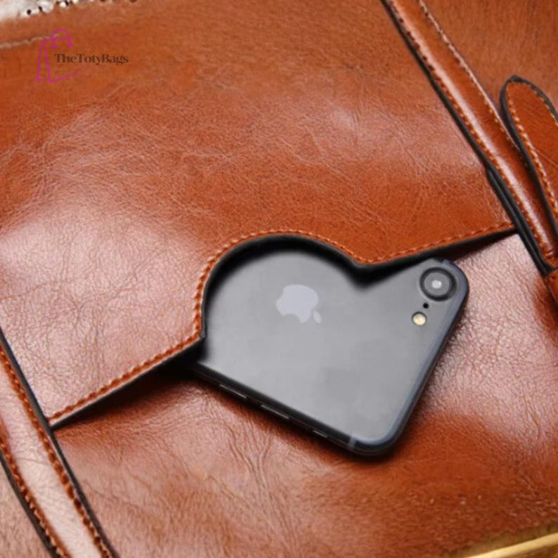 Może przedstawiać: Zbliżenie brązowej sk&oacute;rzanej torebki z kieszenią na czarny smartfon. Torba ma teksturowaną powierzchnię i widoczne szwy. Logo TotyBags znajduje się w lewym g&oacute;rnym rogu.