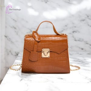 May include: A brown, textured handbag with a top handle and gold chain strap. The bag features a gold clasp closure and a small, decorative flap. The bag is made of a material that resembles crocodile skin.