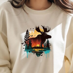 May include: A cream-colored sweatshirt with a graphic print of a moose standing in a forest with a sunset in the background. The moose is black and the forest is green and brown. The sunset is orange and yellow. The graphic is surrounded by a blue and turquoise drip effect.