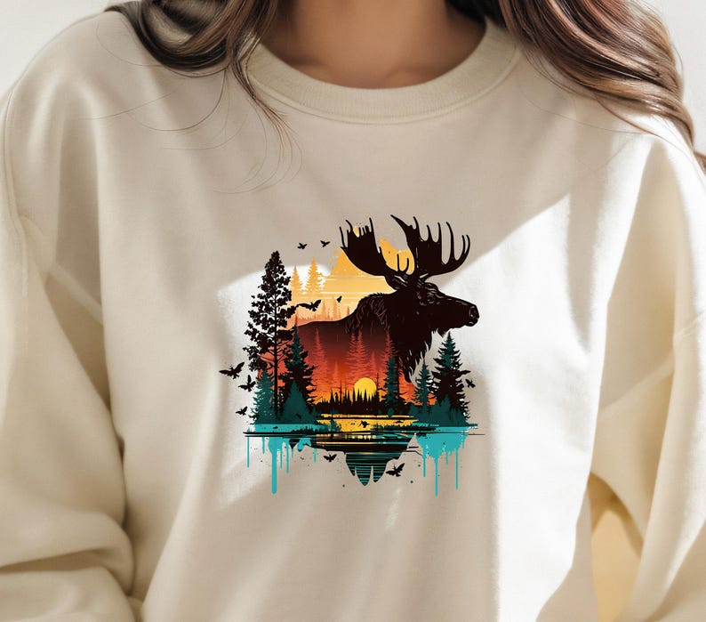 May include: A cream-colored sweatshirt with a graphic print of a moose standing in a forest with a sunset in the background. The moose is black and the forest is green and brown. The sunset is orange and yellow. The graphic is surrounded by a blue and turquoise drip effect.