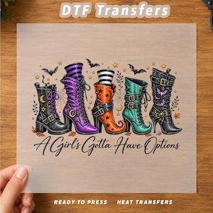 May include: A DTF transfer sheet featuring five distinct boots in black, purple, orange, and teal, decorated with stars, moons, and bats. The text A Girl's Gotta Have Options is printed below the boots. The sheet is labeled DTF Transfers.
