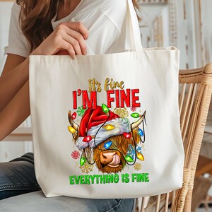 May include: Cream-colored tote bag featuring a Highland cow wearing a Santa hat and Christmas lights. The text "It's fine, I'm fine, Everything is fine" is printed in red, gold, and green. A festive holiday-themed accessory.