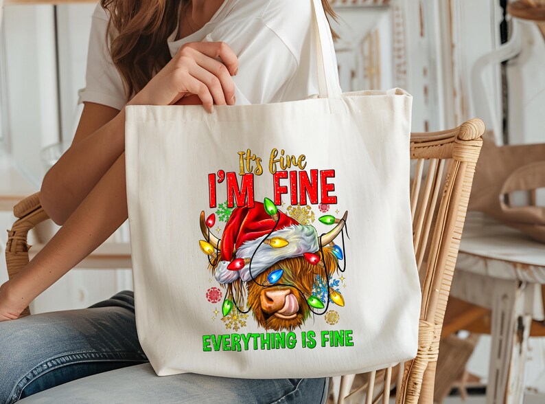 May include: Cream-colored tote bag featuring a Highland cow wearing a Santa hat and Christmas lights. The text "It's fine, I'm fine, Everything is fine" is printed in red, gold, and green. A festive holiday-themed accessory.