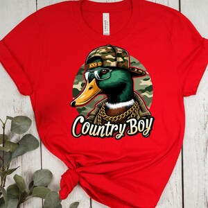 May include: Red t-shirt featuring a graphic of a duck wearing a camouflage hat, sunglasses, and a gold chain. The text "Country Boy" is printed below the duck.