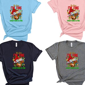 May include: Four t-shirts in light blue, pink, navy, and gray, each featuring a graphic of a Highland cow wearing a Santa hat and Christmas lights. The text reads "It's fine, I'm fine, everything is fine."