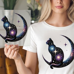 May include: A black cat silhouette with a starry galaxy design sits on a crescent moon. The artwork features a celestial theme with purple, blue, and white colors. The design is on a clear square and a white t-shirt.