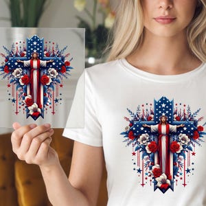 May include: A white t-shirt with a red, white, and blue cross design. The cross is decorated with flowers and stars. A figure of Jesus is on the cross with his arms outstretched.