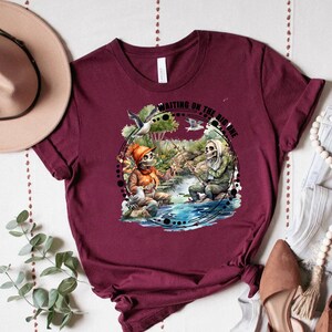 K&ouml;nnte beinhalten: Ein maroonfarbenes T-Shirt mit einem Motiv von zwei Skeletten beim Angeln mit dem Text "Waiting on the big one".