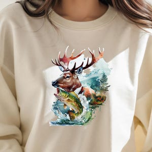 May include: Cream-coloured sweatshirt with a watercolour design of a deer with large antlers, a fish, and a mountain landscape. The artwork includes shades of brown, green, blue, and white. The design is centred on the front of the sweatshirt.