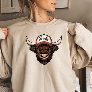 May include: Beige sweatshirt featuring a graphic of a Highland cow wearing a baseball cap with the word "Howdy" in a script font. The cow is brown with long horns and a detailed face. The cap is orange and white.
