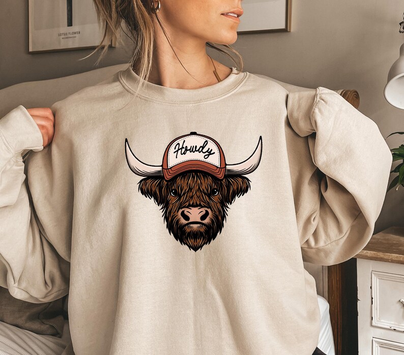 May include: Beige sweatshirt featuring a graphic of a Highland cow wearing a baseball cap with the word "Howdy" in a script font. The cow is brown with long horns and a detailed face. The cap is orange and white.