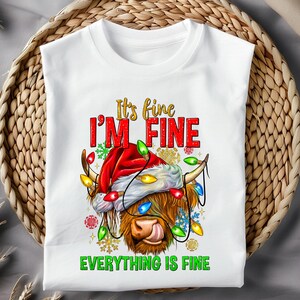 May include: White t-shirt with a graphic of a Highland cow wearing a Santa hat and Christmas lights. The text reads "It's fine, I'm fine, Everything is fine" in red and green. The shirt is displayed on a woven basket.