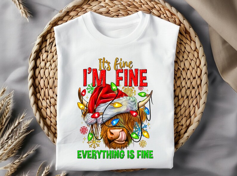 May include: White t-shirt with a graphic of a Highland cow wearing a Santa hat and Christmas lights. The text reads "It's fine, I'm fine, Everything is fine" in red and green. The shirt is displayed on a woven basket.