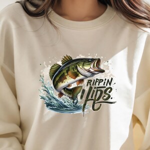 Może przedstawiać: Kremowa bluza z nadrukiem dużego okonia, kt&oacute;ry wyskakuje z wody. Nadruk zawiera tekst "Rippin' Lips".