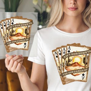 May include: A white t-shirt with a graphic of four playing cards featuring a desert scene with the text "Ain't goin' down til the sun come up".